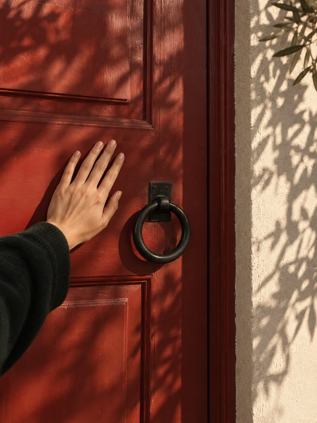 A hand resting on a red door beside a pale plaster wall.